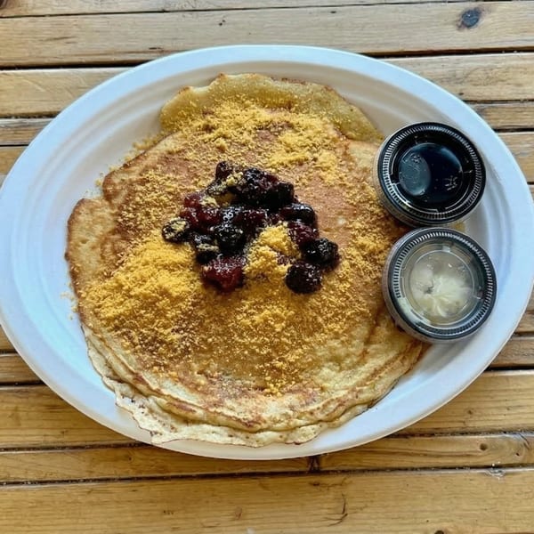 A plate of pancakes topped with yellow powder and dark fruit compote, with two sauce cups.