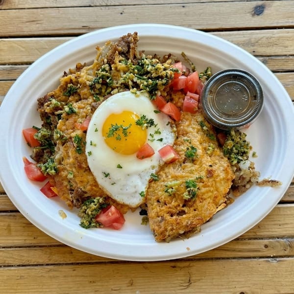 A breakfast plate with fried egg, hash browns, tomatoes, and a side of sauce on a wooden table.