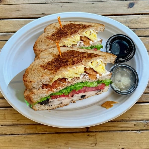 A grilled sandwich with scrambled eggs, bacon, lettuce, tomato, and chicken on a white plate.