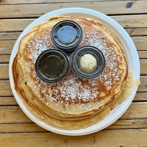 A stack of powdered sugar pancakes with syrup and butter on a white plate atop a wooden table.