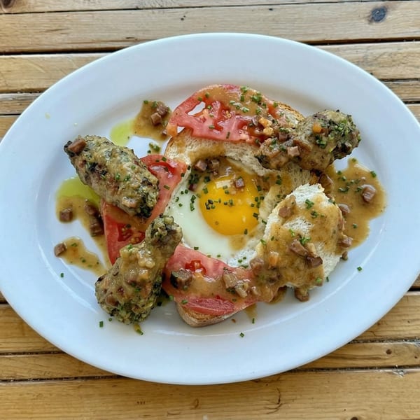 A white plate holds toast with tomato slices, a fried egg, and sausage links covered in gravy.